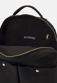 Mochila negra con una cremallera dorada, tela suave, un bolsillo interior con cremallera y una etiqueta etiquetada como "ANNA FIELD". Diseño minimalista.