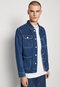 Giacca di jeans blu scuro, con colletto, bottoni frontali e quattro tasche. Tessuto di peso medio, con una texture e dettagli di cucitura visibili.