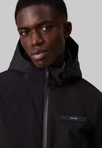 Giacca nera con cappuccio realizzata in materiale liscio e leggero. Presenta una chiusura con zip e un piccolo etichetta con il marchio sul lato sinistro del petto.