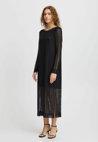 Rochie neagră cu mâneci lungi din țesătură transparentă cu dungi, având linii verticale și o croială dreaptă, terminată cu o lungime sub genunchi și detalii cu fante.