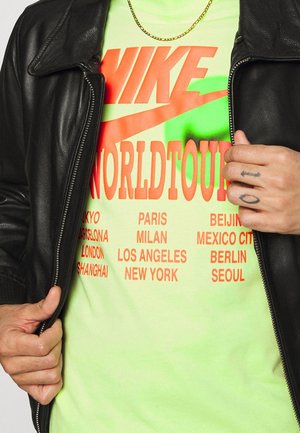 Svart läderjacka över en neongrön t-shirt som visar orange text med "NIKE WORLDTOUR" och stadnamn i fetstil, uppradade vertikalt.