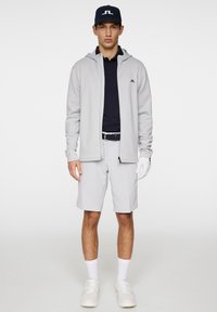 Graue Zip-Jacke über einem dunklen Poloshirt, kombiniert mit hellgrauen Shorts. Weiße Sneaker und eine dunkelblaue Kappe mit einem Logo. Golfhandschuh an der linken Hand.