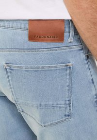 Jeans de mezclilla azul claro con una etiqueta de cuero marrón grabada con "FACONNABLE" en la cintura, que cuentan con bolsillos traseros estándar y detalles de costura.