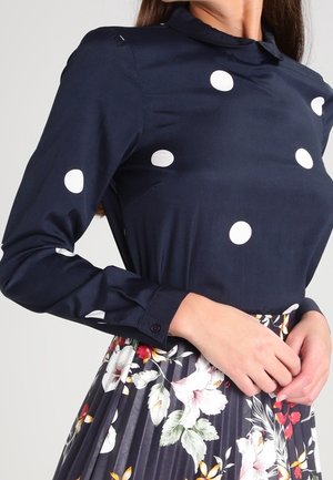 Kobieta w granatowej bluzce w białe kropki i plisowanej spódnicy w kwiaty, trzymająca ręce na talii.