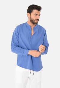 Camicia blu a maniche lunghe con colletto alla cinese, bottoni sul collo e vestibilità rilassata. Tessuto liscio con un aspetto leggermente strutturato.