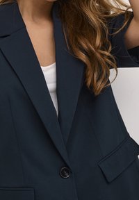 Blazer azul marino con solapa de muesca, cierre de un solo botón y dos bolsillos frontales. Tejido suave con un ajuste a medida, mangas cortas.