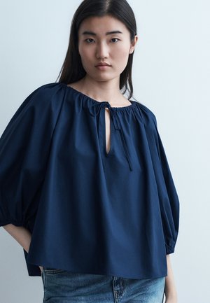 Noor naine, kes kannab lahtist tumelilla pluusi lipsukaelusega ja 3/4 pikkuste volangvarrukatega, kombineeritud siniste teksadega, ühevärvilise heleda tausta ees.