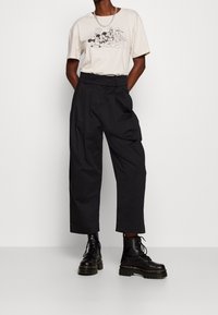 Beige t-shirt med grafisk tryck som föreställer tecknad karaktärer. Svarta vida byxor och svarta snöriga ankelskor med tjocka sulor.