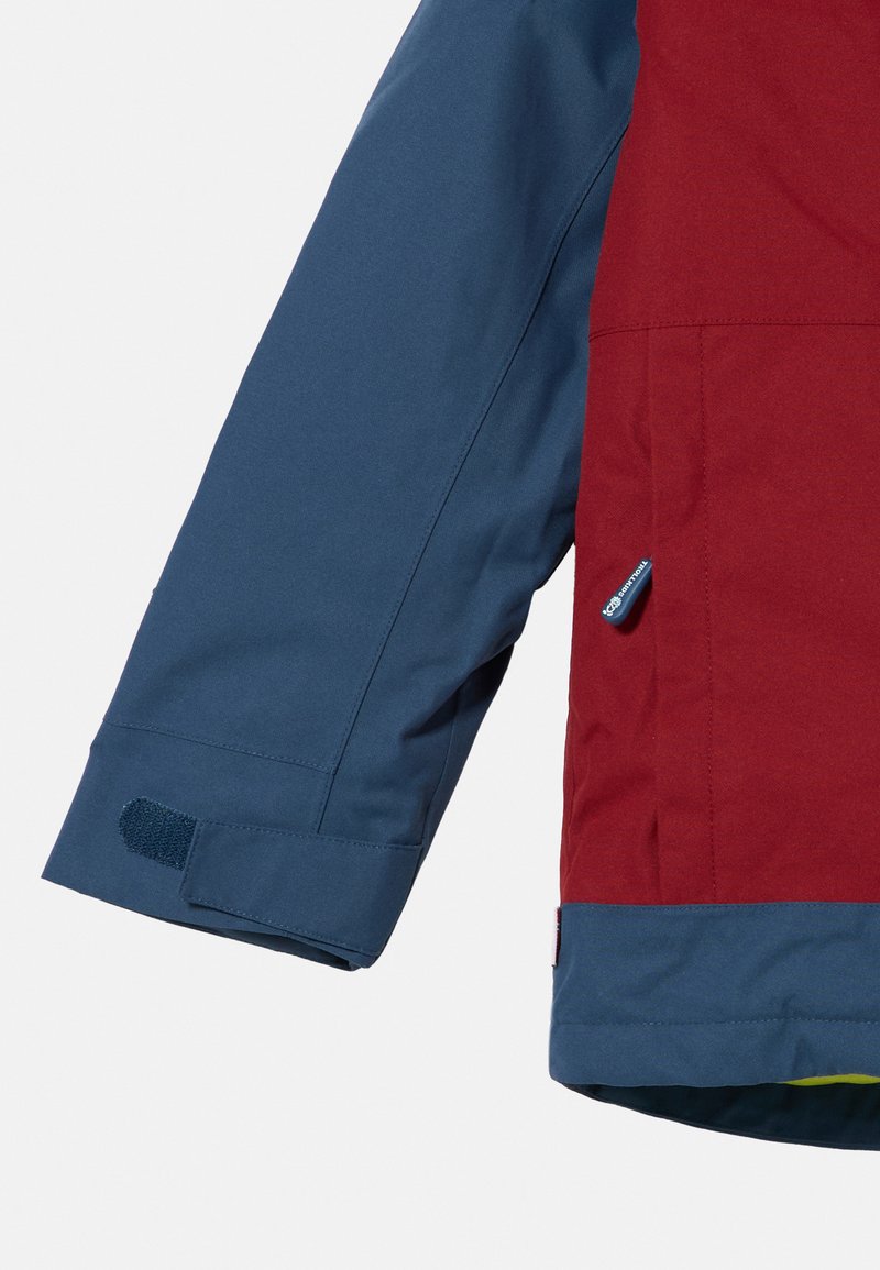 Manga de chaqueta impermeable roja y azul que incluye un ajustador de puño de Velcro, textura suave y paneles de color contrastante. Incluye logo de identificación.