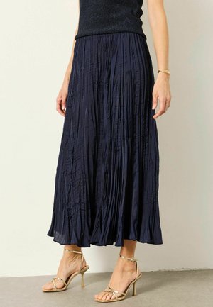 Frau trägt einen marineblauen plissierten Midirock und goldene, hochhackige Riemchensandalen und steht auf einem grauen Boden.