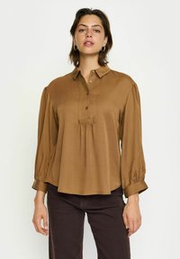 Pruun nööpidega pluus, millel on pikad puff-sleeved ja plekitud esidetail. Valmistatud siledast kangast, klassikalise kraega ja peene läikega.