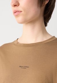 Rudas medvilninis marškinėliai su apvalia iškerpte, turintis mažą juodą logotipą ant krūtinės ir lygią, vienodą tekstūrą.
