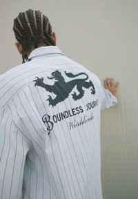 Camicia bianca a righe verticali con una grafica di un leone nero e il testo "Boundless Journey Worldwide" sul retro; tessuto leggero; vestibilità comoda.