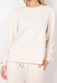 Mujer vestida con una blusa de manga larga color crema y pantalones de cordón a juego, de pie con una mano en el bolsillo frente a un fondo liso.