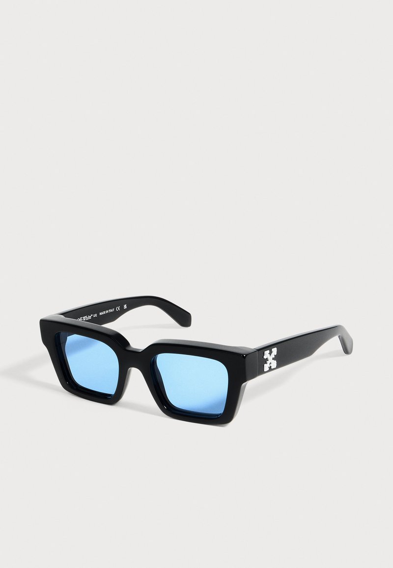 Lunettes de soleil rectangulaires noires avec des montures épaisses et des verres bleu clair, présentant un logo croix blanche sur la branche.