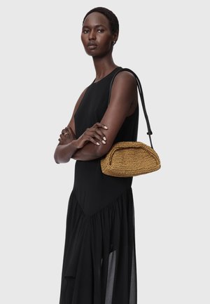 Γυναίκα με μαύρο αμάνικο φόρεμα και σταυρωμένα χέρια, κρατώντας μια μικρή πλεκτή τσάντα ταμπά με μαύρο λουράκι στον ώμο.