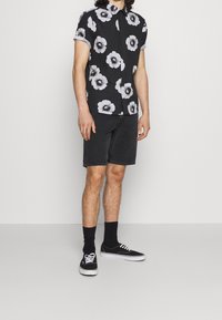 Schwarzes Kurzarm-Hemd mit Knöpfen und großen weißen Blumenmotiven, getragen mit schwarzen Denim-Shorts und schwarzen Sneakern mit weißen Sohlen.