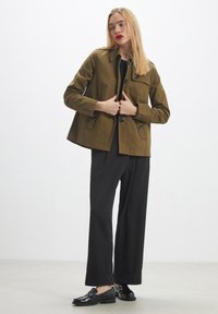 Donna con capelli biondi lisci che indossa una giacca oliva, pantaloni neri a gamba larga e mocassini neri, in piedi contro uno sfondo chiaro e semplice.