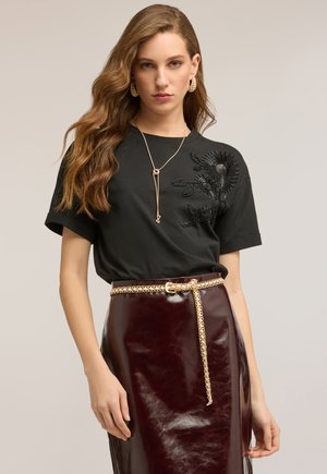 Kobieta o długich falowanych włosach, ubrana w czarną, zdobioną koralikami floralną bluzkę, złoty łańcuszek na szyi oraz kolczyki, a także błyszczącą ciemną bordową spódnicę z paskiem ze złotego łańcuszka.