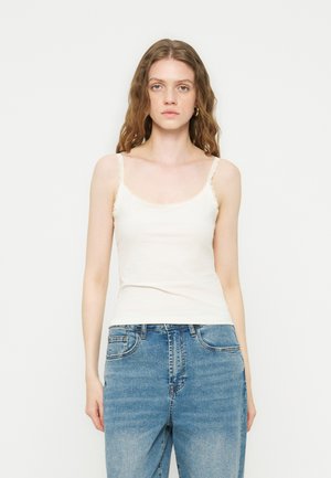 Jeune femme aux cheveux ondulés portant un haut caraco blanc orné de dentelle et un jean en denim bleu sur un fond blanc uni.