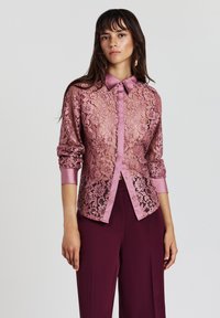Junge Frau in einer durchsichtigen rosa Spitzenbluse mit aufgerollten Ärmeln und dunkelvioletten, weiten Hosen, die vor einem schlichten Hintergrund steht.