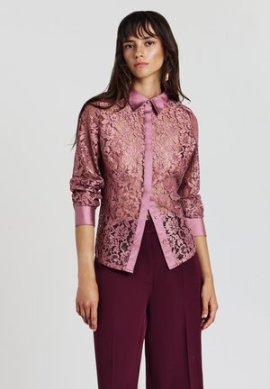 Giovane donna che indossa una blusa in pizzo rosa trasparente con maniche arrotolate e pantaloni larghi viola scuro, in piedi davanti a uno sfondo neutro.