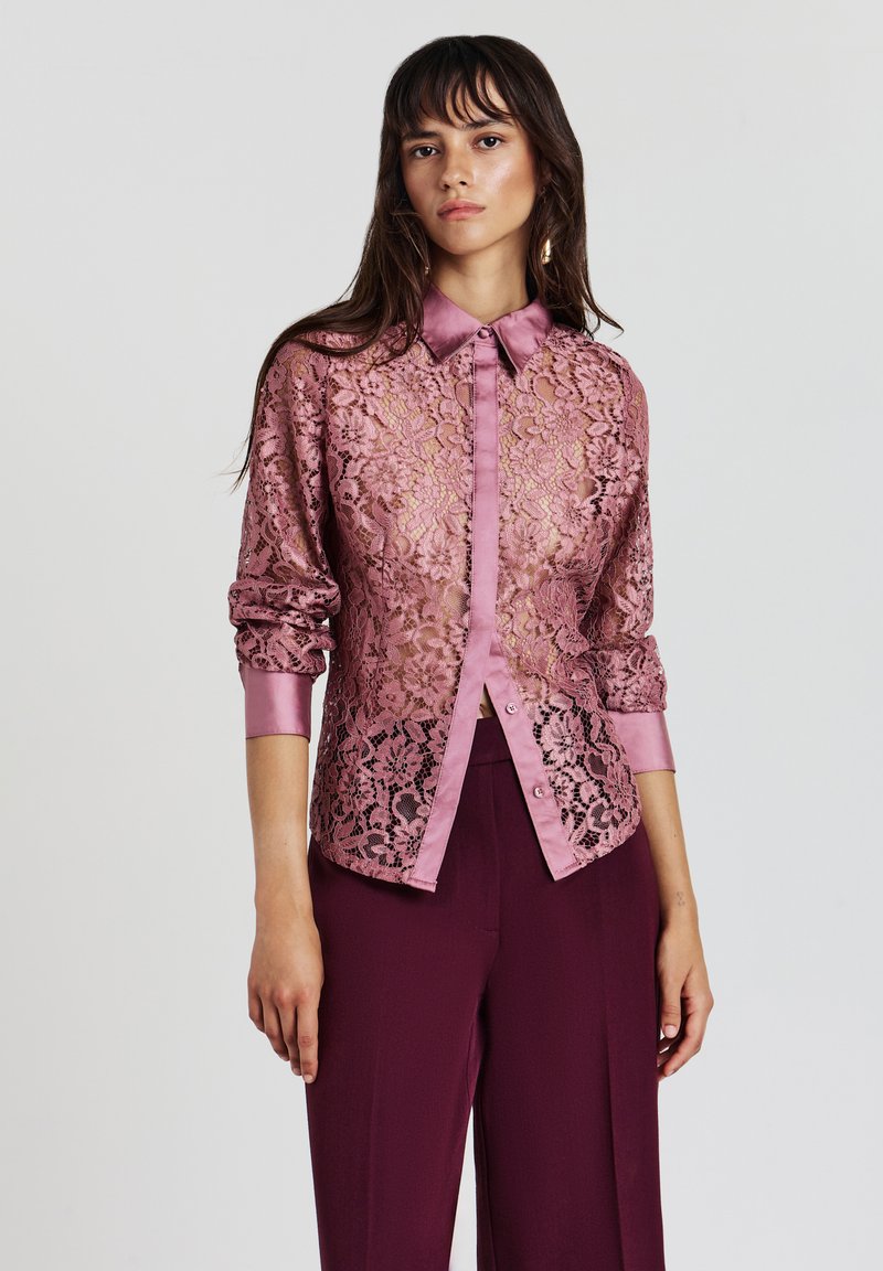 Junge Frau in einer durchsichtigen rosa Spitzenbluse mit aufgerollten Ärmeln und dunkelvioletten, weiten Hosen, die vor einem schlichten Hintergrund steht.