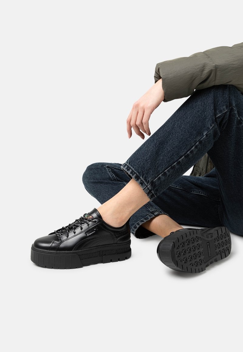 Personne portant des baskets à plateforme noires et un jean foncé, assise avec une jambe croisée sur l'autre sur un fond neutre.