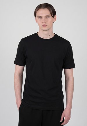 Ung mand med glat mørkt hår iført en simpel sort kortærmet T-shirt, stående med den ene hånd i lommen, neutral grå baggrund.