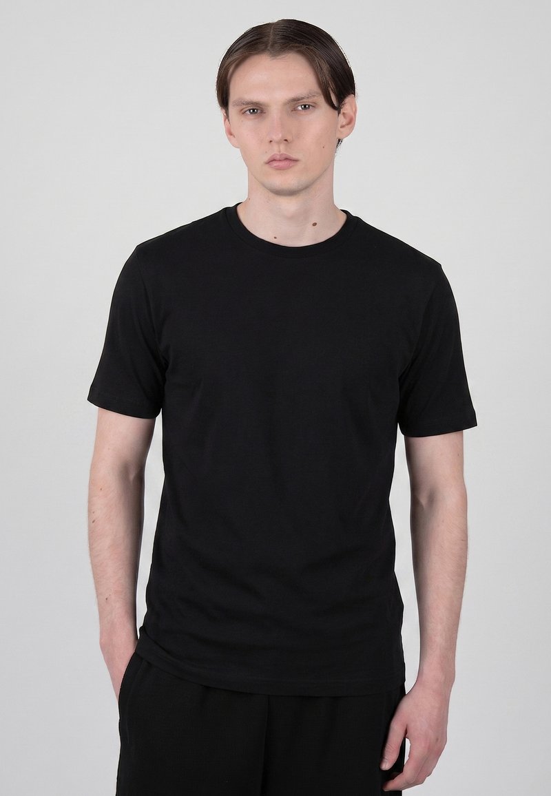 Jeune homme aux cheveux raides et foncés, portant un T-shirt noir uni à manches courtes, debout avec une main dans la poche, fond gris neutre.
