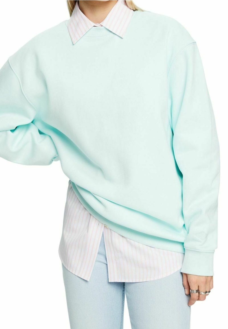 Sudadera de gran tamaño en azul claro con una textura suave, superpuesta sobre una camisa de botones de rayas en rosa pálido y blanco. Ajuste casual, mangas largas.