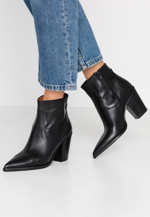 Person trägt blaue, gekürzte Jeans und schwarze, spitz zulaufende Leder-Stiefeletten mit Blockabsätzen und steht auf einem weißen Boden.