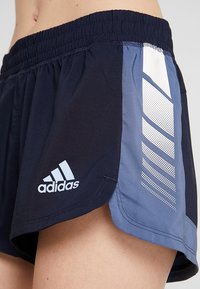 Tummansiniset ja vaalensiniset urheilusortsit, joissa on joustava vyötärö, Adidas-logo ja kulmastriippumainen kuosi, joka on tehty rei'itetystä materiaalista.