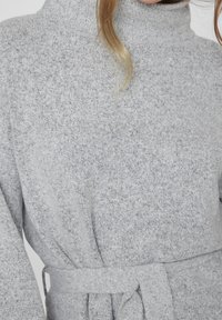 Gros plan d'une personne portant un pull à col roulé en tricot gris clair avec une ceinture assortie nouée à la taille.