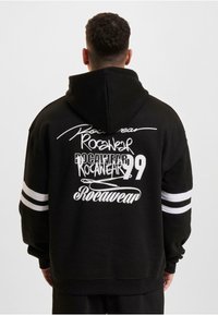 Crna kapuljača s bijelim grafičkim tekstom, uključujući "Rocawear" i broj "99." Dizajnirana sa dvije bijele pruge na svakoj rukavu.