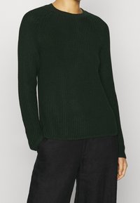 Maglione lavorato a maglia verde scuro con maniche lunghe, texture a costine e scollatura a girocollo; abbinato a pantaloni neri a gamba larga.