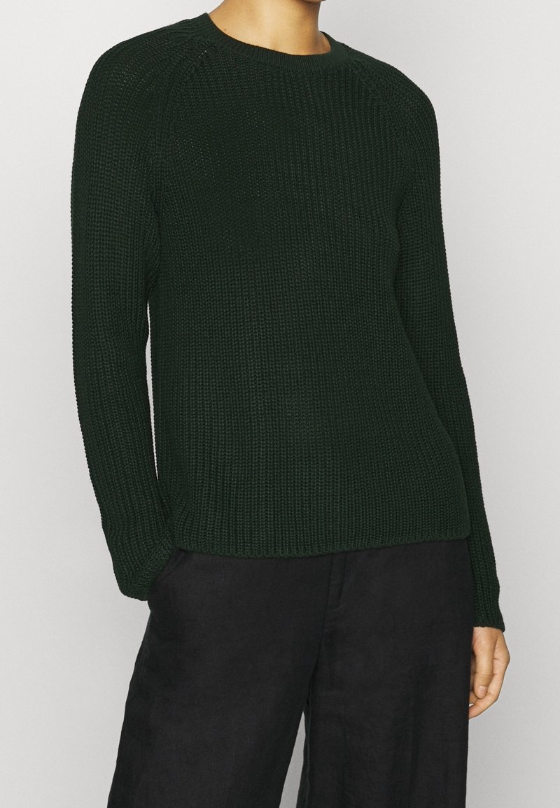 Maglione lavorato a maglia verde scuro con maniche lunghe, texture a costine e scollatura a girocollo; abbinato a pantaloni neri a gamba larga.
