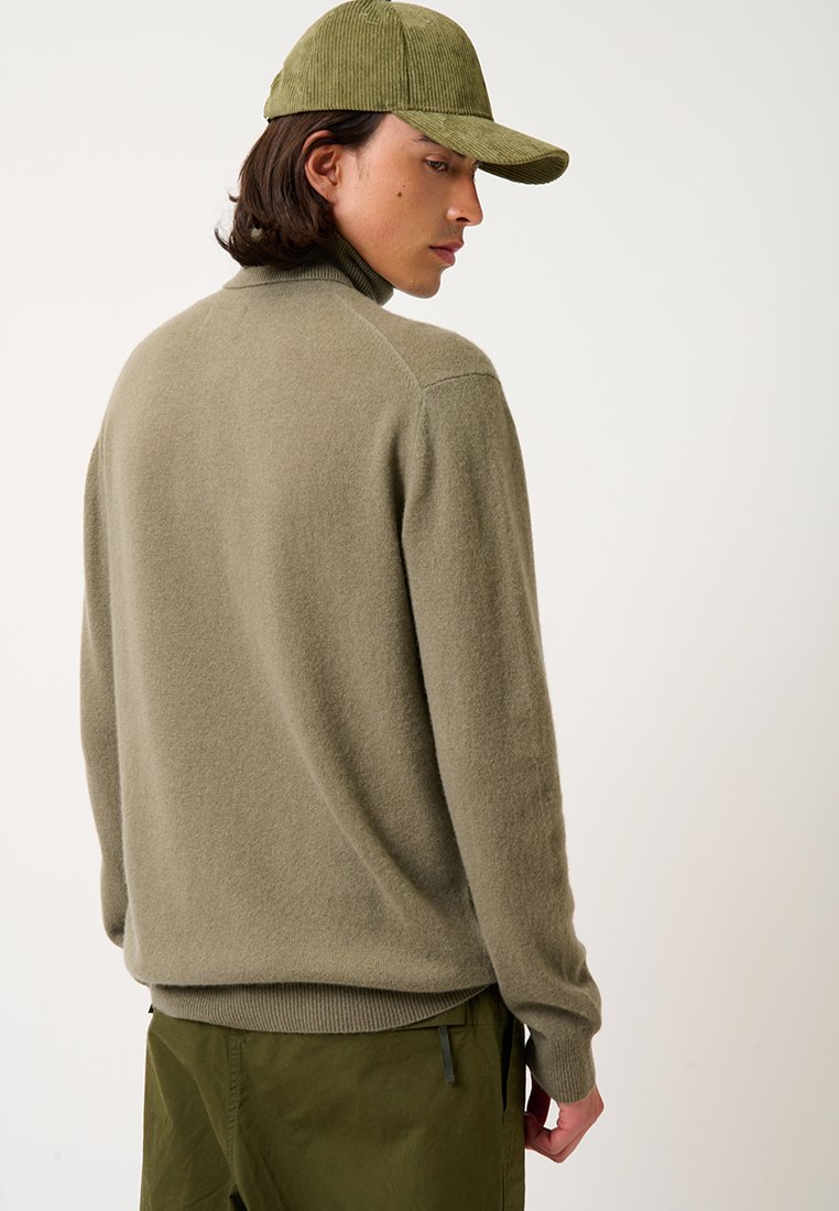 Olivgrüner Wollpullover mit hohem Kragen, geripptem Saum und langen Ärmeln. Kombiniert mit einer passenden grünen Cordmütze.