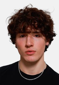 Nærbillede af en person med krøllet mørkebrunt hår, iført en sort t-shirt og en sølvfarvet snoet kædedyse. Neutral baggrund.