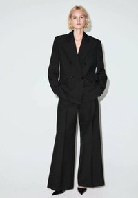 Czarna dwurzędowa marynarka o dopasowanym fasonie, zestawiona z czarnymi spodniami o szerokich nogawkach. Gładki materiał z czystymi liniami i wyraźnym krawiectwem.