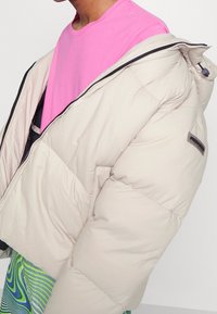 Beige puffer jacket with a zip, featuring a high collar and side pockets, worn over a pink shirt, showcasing a smooth texture and minimal design.
