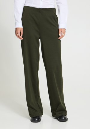 Persoon die olijfgroene wijde broek, zwarte schoenen en een wit shirt met lange mouwen draagt, staand tegen een effen achtergrond.