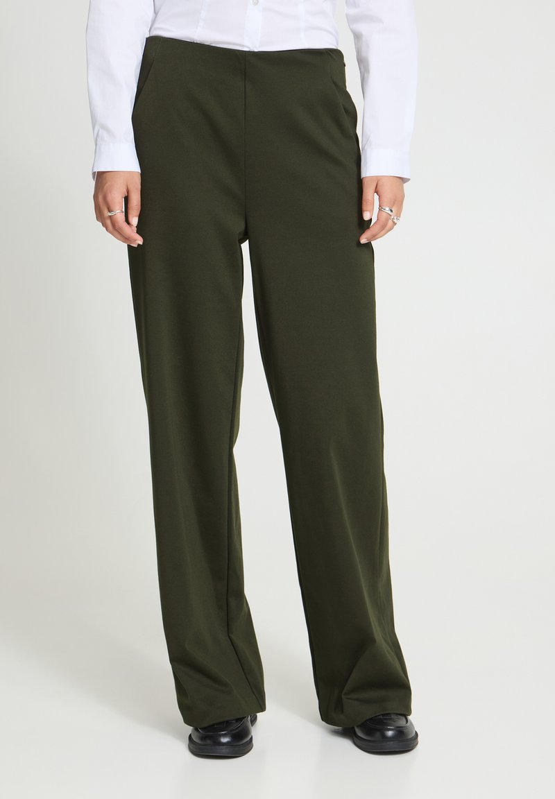 Personne portant un pantalon large vert olive, des chaussures noires et une chemise blanche à manches longues, debout devant un fond uni.