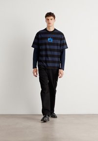 Black and navy striped oversized t-shirt with blue graphic on chest, worn over a long-sleeve top and paired with black cargo pants and shoes.