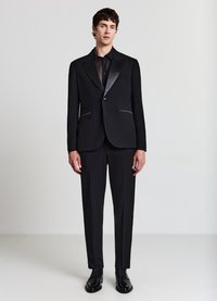 Schwarzer Anzug mit Satin-Spitzenrevers, zwei vorderen Taschen und passenden Hosen. Kombiniert mit einem schwarzen Hemd zum Knöpfen und schwarzen Schuhen.