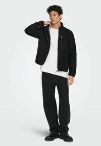 Mladić u crnoj flis jakni preko bijele majice, s crnim hlačama i crnim cipelama, stoji protiv jednostavne svijetle pozadine.