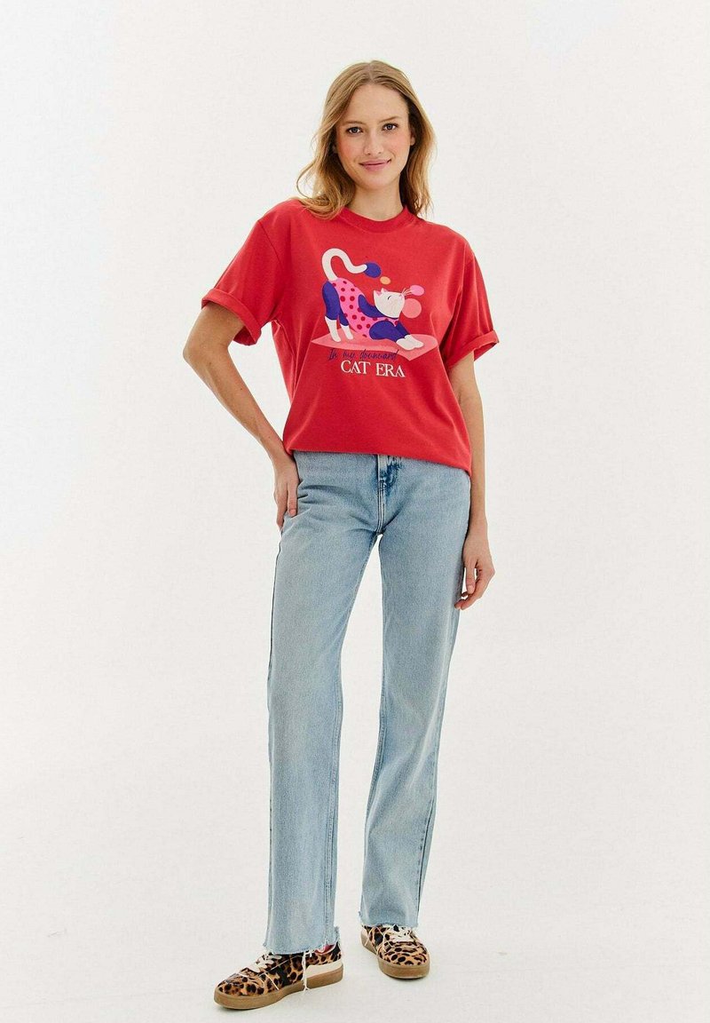 Czerwona bawełniana koszulka z kolorowym nadrukiem kota, w połączeniu z jasnoniebieskimi dżinsami i butami w panterkę, ustawiona na tle białym.