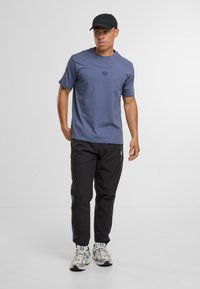 Niebieska koszulka z krótkim rękawem z okrągłym logo, sparowana z czarnymi spodniami z bocznymi paskami i srebrnymi butami sportowymi. Casulowa stylizacja.