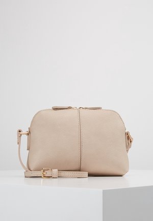 Kleine beige leren crossbodytas met gouden rits en verstelbare band, geplaatst op een wit oppervlak tegen een grijze achtergrond.