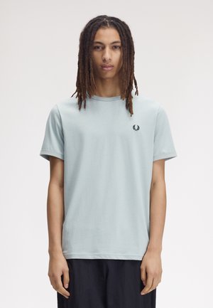 Giovane con lunghi dreadlocks che indossa una maglietta azzurro chiaro con un piccolo emblema nero sul petto, in piedi davanti a uno sfondo semplice.